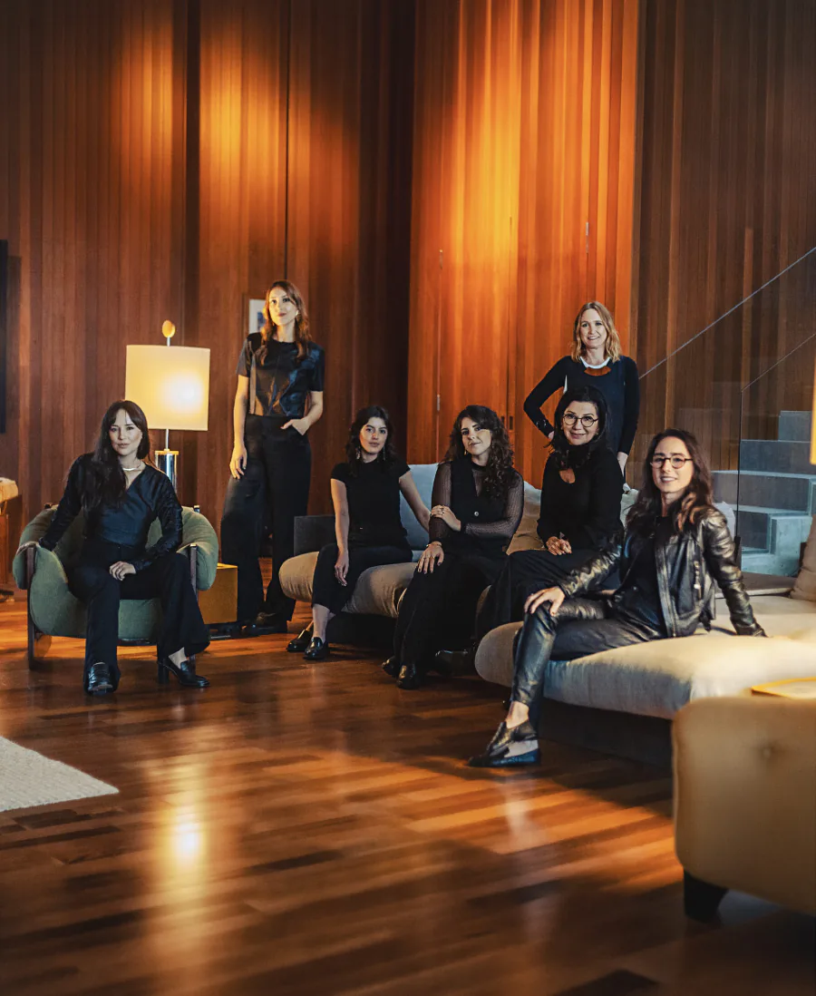 Equipe de sete mulheres reunida num ambiente de madeira, sentadas em poltronas e sofá